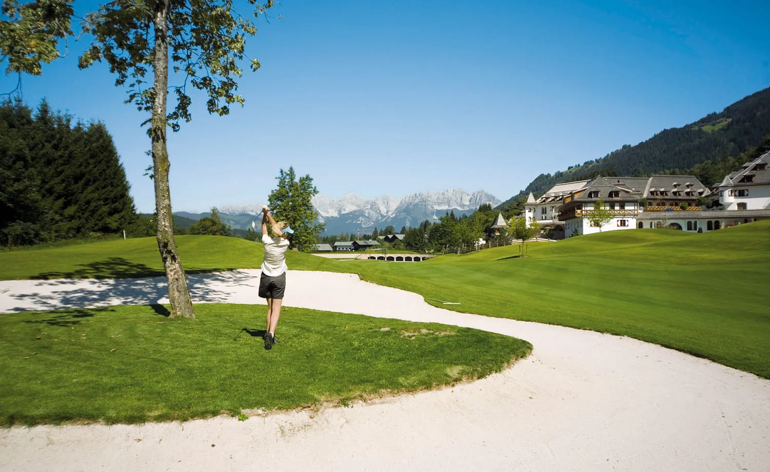 Golferin schlägt neben einem Bunker ab, dahinter ein Hotel und Berge