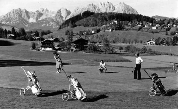Golfer mit Golfwagen auf dem Grün, dahinter Bauernhöfe und Berge
