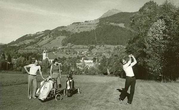 Golfer beim Abschlag, zwei Zuschauer mit Golfwagen, dahinter die Stadt