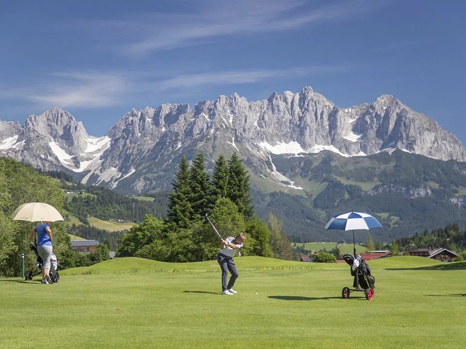 Golfer beim Abschlag vor dem Wilden Kaiser, daneben Trolleys mit Schirmen